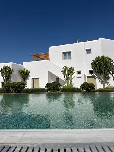 The image depicts a tranquil outdoor scene featuring modern architecture and a serene pool. The subjects are primarily architectural elements: a series of white, multi-story buildings with clean lines and minimalist design. These buildings have rectangular windows and doors, some with wooden shutters or louvers, suggesting a Mediterranean aesthetic. Lush green vegetation, including cactus-like plants and manicured shrubs, is strategically placed around the buildings, creating a harmonious blend of nature and design. A clear, calm pool of water occupies the right side of the frame, reflecting the bright blue sky and the surrounding environment. The water has a gentle ripple, indicating a peaceful atmosphere.
There is no discernible activity or event taking place; the scene is one of stillness and relaxation. The notable details include the stark white of the buildings contrasting with the deep blue of the sky, suggesting a bright, sunny day. The lighting indicates it is daytime, likely midday given the strong shadows cast by the plants and architectural features. The presence of modern, white structures and arid-climate plants, along with the clear blue sky, strongly hints at a location in a sunny, warm climate, consistent with the provided location context of Agios Ioannis Diakoftis, Greece. The overall emotion conveyed is one of peace, luxury, and a quiet escape. No visible text is present in the image.