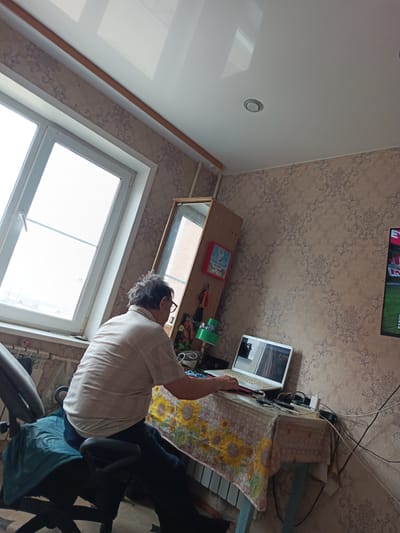 An elderly male with graying hair, wearing glasses, a light-colored checkered shirt, and dark trousers, is seated at a desk in what appears to be a room in Gusinoozyorsk, Russia. He is using a white laptop positioned on a table covered with a tablecloth featuring a sunflower pattern. The table is cluttered with various items, including power cords, a white power adapter, and a dark object. To the left of the laptop, a green desk lamp is visible. Behind the table, a tall, narrow mirror-like object stands vertically. The room has wallpaper with a damask pattern and a white, glossy ceiling with recessed lighting. A large window with a white frame dominates the left side of the image, showing a bright, overcast sky outside. A dark office chair with a teal cover on the seat is where the man is sitting. To the right of the scene, a television screen displaying an image with text and colors is partially visible. The overall impression is that of a domestic setting, likely a home office or living space.