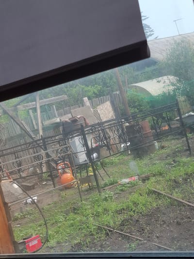 An outdoor, overcast scene captures a yard in Mariinsk, Russia, viewed through a window. A dark, horizontal blind or shade obscures the top portion of the image. The yard itself is cluttered with various items. A black metal fence with decorative scrollwork runs across the mid-ground. To the left of the fence, an orange air compressor and a red welding machine are visible on the ground, with a black cable extending from the welding machine. Behind the fence, a brown barrel stands next to a white sheet or tarp. The ground is covered with grass and dirt. In the background, a greenhouse-like structure with a curved, translucent roof is situated amongst dense green foliage. A wooden fence and some construction materials are also present. The overall impression is one of a somewhat unkempt outdoor working or storage area.