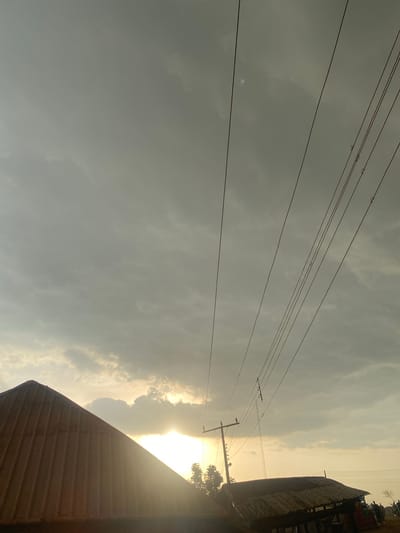This image depicts a dusky sky over Jere South, Nigeria, with a prominent power line stretching diagonally across the frame. The setting sun casts a warm glow, partially obscured by clouds, near the horizon. In the foreground, a section of a corrugated metal roof is visible on the left, its metallic sheen catching the light. To the right, another, more dilapidated corrugated structure is present, its roof appearing weathered. A utility pole supporting the power lines stands between the two structures. The environment appears rural or semi-rural, with some trees visible on the left and a few silhouetted figures standing in the distance on the right, suggesting human presence. The overall mood is serene and somewhat atmospheric due to the dramatic lighting.