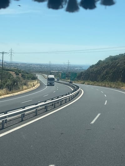 A white semi-trailer truck is traveling on a highway. The truck is in the right lane. The highway is curving to the right. There are trees and hills on the right side of the highway. There are power lines and a town in the background. The sky is blue and clear. A bird is visible in the sky. The image appears to be taken from a vehicle traveling on the highway. This is a view of the Attiki Odos highway near Avra, Greece.