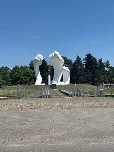 The image depicts a monument in the city of Poti, Georgia. The monument features two large, abstract white sculptures resembling waves or unfurling petals. Centered between these sculptures is a smaller, golden statue of a man standing on a pedestal. The monument is situated in an open area with a gravel path leading towards it, bordered by a metal fence. Behind the monument, there is a dense line of dark green trees, suggesting a park or natural setting. The sky above is clear and blue with a few faint clouds. The foreground shows a paved area, likely a road or parking lot, with loose gravel. The overall scene suggests a public monument or memorial site.