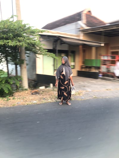 In the city of Glagah Arum, Indonesia, a woman wearing a grey hijab and a black dress with a tan leaf pattern walks on a paved road. She is carrying a translucent plastic bag in her right hand. To her left is a tree with green leaves and a light-colored pole. Behind her, a building with a brown, tiled roof and beige walls is visible. The scene is slightly blurred, suggesting movement.