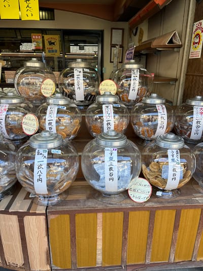 The image shows a variety of glass jars filled with different types of snacks, likely Japanese sweets, displayed on a wooden shelf. The jars are arranged in three rows and are labeled with Japanese characters. The jars are located inside a shop. The image appears to have been taken from a slightly elevated angle, looking down onto the jars. The background is a wooden wall with a yellow color. The photo appears to be taken in front of a store in Katsushika City, Japan. The coordinates 35.758, 139.877 place this scene in Tokyo, Japan.