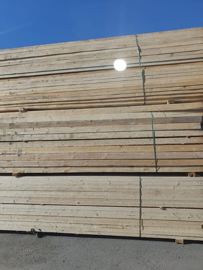 This media file captures a close-up, high-angle view of a large stack of lumber. The lumber planks are arranged vertically, side-by-side, and bound together with green strapping. The texture of the wood is rough and weathered, with visible grain patterns and occasional knots. The lighting suggests it's daytime, with a bright light source causing a strong glare on some of the planks. In the upper left portion of the image, a clear blue sky is visible, indicating fair weather. A portion of a gray, asphalt-like surface is present on the right side of the frame, potentially representing the ground or a road.
Notable details include a bright circular glare, possibly from the sun or a light source, shining on the wood. Faint shadows are cast by the strapping onto the planks. There is a small mark with the numeral "110" written on one of the planks. The scene appears to be an outdoor storage area for construction materials, possibly at a lumber yard or worksite. The overall impression is one of a practical, functional setting. There are no people or animals visible in the image. The time of day is likely midday or late afternoon due to the intensity and angle of the light.