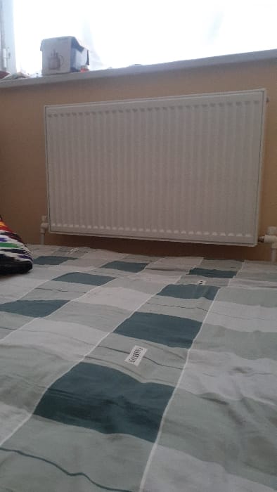 A radiator hangs vertically on a wall, adjacent to a window and a checkered bedspread. The radiator is white and features a standard design with multiple vertical panels. A small white box is visible on the left, near the window. The bedspread exhibits a gray and teal checkered pattern, with a white tag reading "FASHION" sewn onto it.
