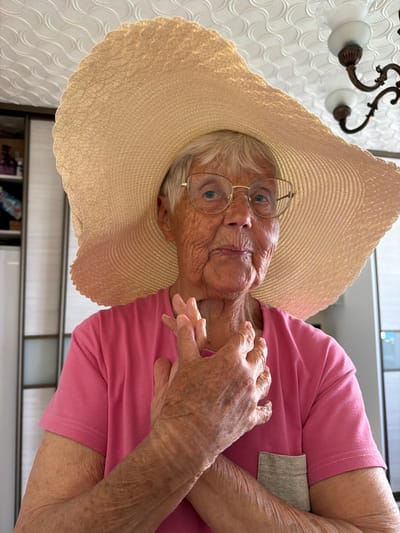 An elderly woman with wrinkles and blonde hair, wearing a large straw hat and round gold-rimmed glasses, is looking directly at the camera. She is wearing a pink short-sleeved shirt with a pocket. Her arms are crossed in front of her. The background includes a patterned white wall with a decorative light fixture and a partial view of what appears to be a shelf or cabinet. The location and city of Valozhyn, Belarus, cannot be definitively determined from this image alone.