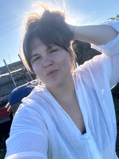 This is a close-up, rotated selfie of a woman in Kraishevo, Russia. She is wearing a loose-fitting white linen shirt. The sun is bright, casting a strong backlight that illuminates her hair and the fabric of her shirt, creating a hazy, ethereal effect. The sky is clear and blue. In the background, there are glimpses of what appears to be an outdoor setting with some weathered wooden structures, possibly a fence or shed, and vegetation. A blue tarp is also visible behind her. The woman's expression is relaxed, with her head tilted back and her hand resting on her head. The overall mood is serene and summery, suggesting a peaceful moment outdoors during the daytime, likely late afternoon given the warm light. There is no visible text in the image.