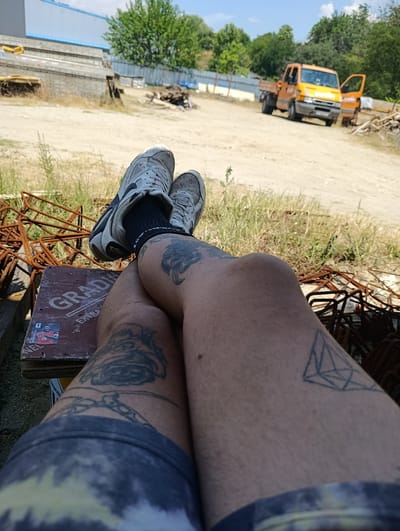 The image depicts a first-person perspective, with the viewer's legs, clad in shorts and athletic shoes, prominently featured in the foreground. The individual is seated on a wooden object, possibly a crate, which is adorned with stickers and text. The legs are tattooed with various designs, including a floral pattern and geometric shapes. Scattered around the immediate area are piles of rusty metal, likely rebar, and dry vegetation.
In the background, a construction or industrial site is visible under a clear blue sky. A bright orange utility truck with its doors open is parked on a dirt path. Further in the distance, a long, light-colored building with a blue roof can be seen, along with trees and fencing. The overall scene suggests an outdoor, possibly rural or industrial, environment. The presence of the truck and building implies a location related to construction or heavy industry. While the image is not definitive, the prompt specifies it is from Struma, Bulgaria.