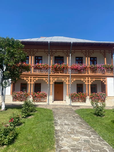 The image shows a two-story building with a white facade and wooden balconies overflowing with red and pink flowers. The balconies are supported by wooden pillars with intricate carvings. There are windows with dark wooden frames on both floors, and a double wooden door in the center of the ground floor. A stone pathway leads up to the entrance, flanked by a green lawn and some rose bushes. A large tree with green leaves is visible on the left side of the building. The sky is clear and blue. This building is located in the village of Filioara, Romania.