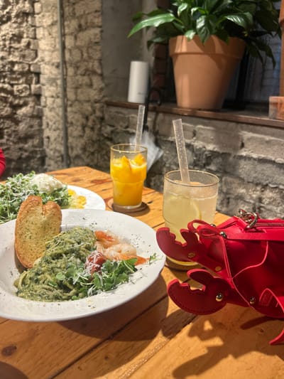 The image captures a dining table scene within a restaurant in 마포구, South Korea. The wooden table surface is laden with food and beverages. A prominent white bowl in the foreground contains green pesto pasta, accompanied by cooked shrimp, sliced cherry tomatoes, fresh microgreens, and grated cheese, along with a slice of toasted garlic bread. To the left, a second white plate holds a green salad topped with a white cheese, possibly burrata, and scattered yellow corn kernels.
Two clear glasses, each containing a clear plastic straw and resting on a brown coaster, are positioned near the center. The left glass holds an orange-colored beverage with fruit slices, while the right glass contains a light yellow beverage with lemon slices. A red, crab-shaped purse with gold-colored stud details is present on the table to the right.
The background reveals a textured stone or brick wall. A shelf runs along this wall, supporting a large potted plant in a terracotta pot on the right and a white cylindrical object, possibly a diffuser, on the left. A folded white umbrella is partially visible behind the drinks. A small portion of a red sleeve is discernible on the extreme left edge of the image, indicating the presence of an individual.