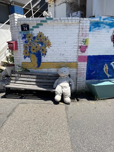 An outdoor scene captured in Jongno 19-gil 74, Jongno-gu, South Korea, under bright daylight. The foreground consists of asphalt pavement, marked by shadows from the objects. A large, light grey teddy bear wearing light-colored overalls is positioned on the pavement, appearing to sit in front of a dark wood-slatted bench with black metal framework, which is situated against a painted brick wall. The bench is unoccupied. The white brick wall behind the bench and teddy bear features two distinct murals: on the left, a large blue vase filled with yellow sunflowers with orange centers and green leaves against a yellow base; on the right, a section of a blue mural depicting a fish, and above it, a smaller pink and yellow section with partial Korean text reading "쉬는". An address plaque displaying "종로19길 74" is affixed to the wall above the sunflower mural. A red metal mailbox is mounted on the wall to the far left. Two white bird figurines are perched on the top edge of the wall above the mailbox. To the left of the bench, a light-colored plant pot contains a small green plant. In the upper background, a white external staircase with dark railings ascends on the left, while corrugated metal siding and a white cooler box branded "Corona" are visible on the right. A turquoise-colored planter or container is present on the far right, near some sparse ground vegetation.