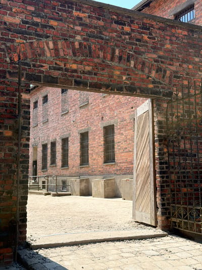 This media file depicts an outdoor scene of old brick buildings with metal-barred windows. The perspective is looking through an archway or opening in a brick structure, revealing a courtyard or alleyway between buildings. The architecture suggests an industrial or historical setting, possibly a former prison or factory. The buildings are constructed with weathered red bricks, and some have a rough, concrete-like exterior. The sky is visible in the upper left corner, indicating it is daytime, likely sunny due to the shadows. The overall mood is somber and perhaps a bit eerie, given the presence of barred windows and the aged, imposing structures. There is a wooden plank structure visible above the opening, and some metal railings on the right side of the scene. No people or text are visible in the image. The location is Oświęcim, Poland.