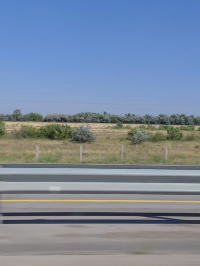 The image captures a roadside scene with a clear blue sky dominating the upper portion. Below the sky, a line of trees and vegetation extends across the horizon. The landscape appears to be dry and grassy with scattered bushes. In the foreground, a gray guardrail with horizontal silver bars runs parallel to the road. A bright yellow line marks the edge of the asphalt road, with dark gray shadows cast by the guardrail and possibly other unseen objects. The scene suggests a travel perspective from a vehicle, given the motion blur on the guardrail and road. The image is set in the city administration of Aqsu, Kazakhstan.