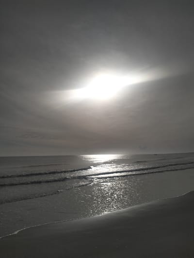 The image shows a serene seascape at Blang Lancang, Indonesia, under a bright, hazy sun. The sun's intense glare dominates the upper portion of the frame, casting a bright reflection on the calm ocean surface. Gentle waves roll towards a sandy shore, with the water shimmering and reflecting the sunlight. In the distance, a small boat is visible on the horizon, adding a sense of scale to the vastness of the ocean. The sky is cloudy, with the clouds diffused by the bright sunlight, creating a soft, ethereal atmosphere. The overall scene is tranquil and evokes a sense of peace and natural beauty.
