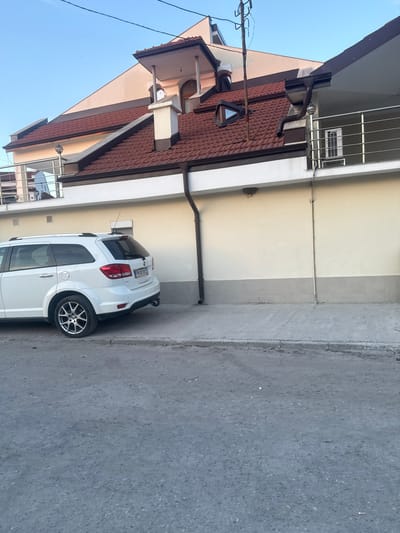 In the city of Benkovski, Bulgaria, a white Dodge Journey SUV is parked on a paved surface adjacent to a building. The building features a light-colored facade, a dark-tiled roof, a balcony with metal railings, and a prominent downspout. The foreground is dominated by a grey, asphalt-like surface, indicating a road or pathway. The sky above is clear and blue, suggesting fair weather. The scene appears to be captured during daylight hours.