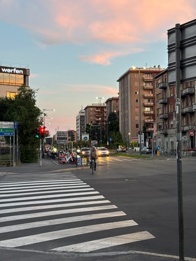 The image depicts a street scene in Sesto San Giovanni, Italy, during what appears to be sunset, with soft pink and orange hues in the sky. In the foreground, a wide zebra crossing dominates the frame, with asphalt road surrounding it. To the left, a building with the prominent white lettering "werfen" is visible, reflecting the sky in its windows. Adjacent to the "werfen" building, a traffic light shows a red signal for the lane. Several motorcycles and scooters are parked near the intersection, and one person is riding a bicycle on the road.
Further into the scene, multi-story residential buildings line the street, exhibiting typical Italian architecture with balconies and various window designs. There are cars on the road, and streetlights are illuminated. Pedestrian signs and street signs are also visible, indicating directions and traffic regulations. The overall environment suggests a moderately busy urban intersection in the late afternoon or early evening.