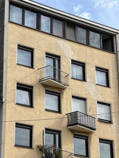 The image shows a section of a multi-story apartment building with a rough, yellow stucco facade. The building features several windows with dark frames, some with vertical blinds visible. There are two small balconies, one with a metal railing and a white floor, and another partially enclosed with a striped fabric screen and decorated with two small blue figures and a flag-like object. A diagonal, lighter-colored stripe runs across the facade of the building. The top of the building has a row of horizontal windows. In the lower left, a gray column is visible. A power line runs horizontally across the middle of the image. In the lower left of the frame, a potted plant with red flowers is visible on a balcony railing. The sky above is partly cloudy. The building is located in Witten, Germany.