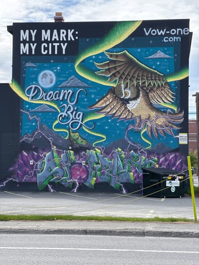 The image displays a vibrant, large-scale mural painted on the side of a dark building, set against a backdrop of a clear, cloudy sky overhead. The mural's central feature is a majestic owl with striking yellow eyes and brown and gold plumage, soaring with outstretched wings against a deep blue night sky filled with white stars and scattered clouds. A prominent full moon is depicted towards the lower left of the mural. Yellow-green ribbons gracefully twine around the owl and across the starry expanse.

Text incorporated into the mural includes "MY MARK: MY CITY" in bold white letters on the left side, and "Dream Big" in elegant script-like font in the center. Vertically oriented text "Vow-one.com" is visible in the upper left corner. The right side of the mural features abstract, geometric designs resembling jagged, purple crystals and green, blocky structures, interconnected by white lightning bolts. Small numerical details, such as "9" and "10," appear on some of these shapes. Another small text "one/CBC" is partially visible at the bottom right of the mural.

In the foreground, a dark green dumpster is placed directly in front of the mural, secured by bright yellow chains. Text on the dumpster reads "Alaska Waste," "4yd," "4012335," and "DO NOT OCCUPY." A sign above the dumpster states "SEED Lab MUSEUM Parking Only." The ground consists of an asphalt parking lot or road, marked with white and yellow lines, and bordered by a strip of green grass and a concrete curb. The scene is illuminated by daylight, providing a clear view of the detailed artwork in Anchorage, United States of America.