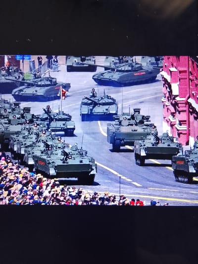 The image depicts a military parade with multiple armored vehicles, including tanks and armored personnel carriers, proceeding down a street lined with spectators. The vehicles are predominantly olive green and appear to be in formation. Several soldiers are visible on top of or inside the vehicles. The spectators are densely packed along the side of the street, many holding up cameras or phones. The architecture on the right side of the image suggests an urban setting, with a red brick building prominently featured. The overall scene is one of a formal military procession. The location is identified as Safonovo, Russia.
