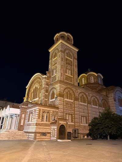 This is a nighttime, low-angle, full-shot photograph of the Church of Saint Nicholas, located in Nea Kallikratia, Greece. The church is constructed of white and tan bricks, with arched windows and a central dome topped with a cross. A tall bell tower stands to the left of the dome, also adorned with intricate brickwork and arched openings. The church is illuminated by artificial lighting, casting a warm glow against the dark night sky. In the foreground, a paved courtyard is visible, with a large tree on the right side of the image. The scene is devoid of people or any observable activity, presenting a serene and architectural view of the religious structure.