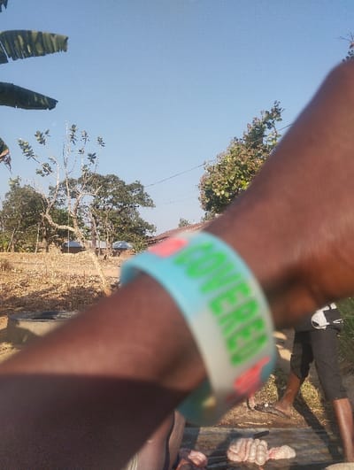 A close-up shows a dark-skinned arm with a light blue bracelet displaying "COVERED" in green letters. In the blurry background, two figures stand near what appears to be animal intestines on a surface. The scene unfolds outdoors under a clear sky, with trees and a small structure visible in the distance. The foreground is dominated by the arm, drawing the viewer's attention to the bracelet and the person wearing it.