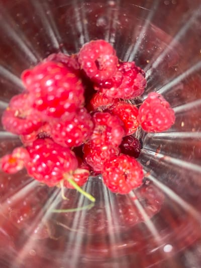 A high-angle, close-up shot shows a collection of red raspberries in a clear, ribbed glass container. The raspberries are plump and juicy, with a few appearing to have a slightly darker, almost black hue, suggesting a variety of ripeness or type. The light glinting off the glass creates streaks and reflections that add depth to the image. One small green stem is visible, attaching to a raspberry in the center-right of the frame. The scene is bright and appears to be indoors, with no indication of the time of day or weather. The smooth, reflective surface of the glass and the texture of the raspberries are the main focus. There is no visible text in the image.