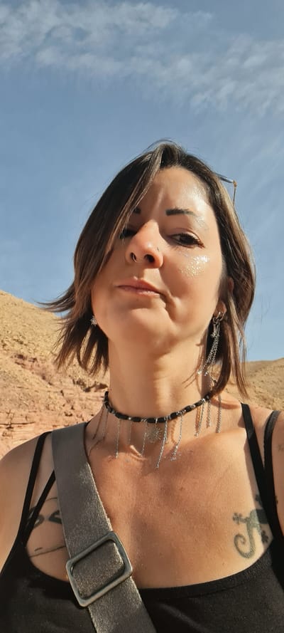 A woman is standing outside, with the sky in the background. She has short brown hair and is wearing a black top, a black beaded necklace with silver charms, and a grey strap over her shoulder. She has a nose ring and shimmering makeup on her face. A tattoo of a lizard is visible on her upper chest. The location is likely near a desert area, as indicated by the sandy-colored ground in the background. The city is Hevel Eilot Regional Council, Israel.