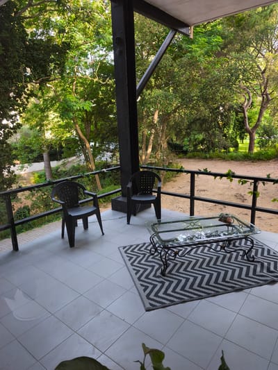 The image shows a covered outdoor patio with a tiled floor, overlooking a wooded area. Two black plastic chairs are placed on the left side of the patio. In front of them, on a black and white chevron rug, is a glass-top coffee table with a dark metal base. The coffee table has a small decorative item on it. A black metal railing runs along the edge of the patio, with some green vines growing on it. The background consists of dense green trees and vegetation, suggesting a natural or park-like setting. The lighting indicates it is daytime, likely late afternoon or early morning due to the warm light. There are no people visible in the image.