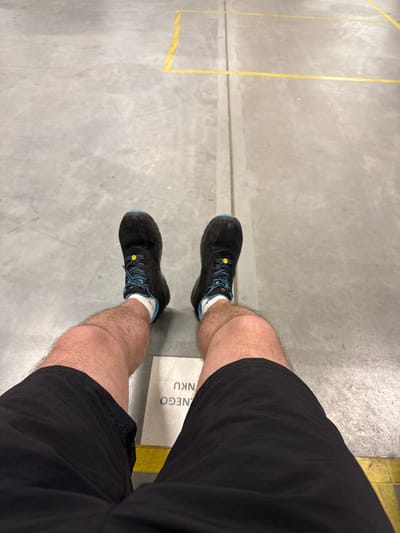A first-person perspective shot shows a person's legs and feet wearing black shorts, white socks, and black and blue athletic shoes. The person is sitting on a concrete floor, with yellow lines painted on the floor in the background. The yellow lines appear to be part of a warehouse or industrial setting. A yellow and white marker with text is visible near the person's feet. The scene suggests a casual or break moment in an industrial or warehouse environment, possibly located in the city of Chociule, Poland, though this is not directly confirmed by visual cues within the image itself.