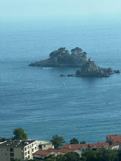 The image displays a vibrant coastal landscape under bright daylight, likely depicting Stupovi, Montenegro, with a prominent rocky islet. The central focus is a rugged, tree-covered island emerging from the clear blue sea. On the island, a small, red-roofed building, possibly a chapel or house, is visible among the greenery. Smaller rock formations extend from the main island into the water.
The expansive sea fills most of the frame, showing visible ripples and textures on its surface, indicating gentle movement. Two small white boats are present on the water, one closer to the islet and another further out in the sea. Along the right side of the image, the coastline features several buildings with distinctive red-tiled roofs, characteristic of Mediterranean architecture. These structures are surrounded by green trees, suggesting a residential or resort area. The sky above is a clear, light blue, without clouds, emphasizing the sunny weather.
No specific human activity is discernible, and no visible text appears in the image. The scene conveys a sense of tranquility and natural beauty, typical of a coastal destination.