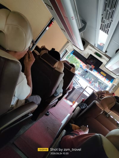 This image depicts the interior of a bus, likely in the city of Gò Dầu, Vietnam, as indicated by the timestamp and the "realme Shot by zsm_travel" watermark. The perspective is from a passenger's seat, looking towards the front of the bus and out the window.
The bus interior features rows of brown and beige upholstered seats, some of which are occupied. In the foreground on the left, a person wearing a light-colored shirt and a beige headrest is visible from behind. Further down the aisle, other passengers are seated. The floor is covered with a red, patterned mat.
The ceiling of the bus has white panels with some decorative red stripes and ventilation grates. A small screen is mounted above the windshield, displaying a digital clock showing "10:36".
Through the windshield, a street scene is visible, suggesting the bus is either stopped or moving slowly. There are buildings, people walking, and vehicles on the street, consistent with an urban or semi-urban environment. The overall lighting suggests it is daytime.
The watermark "realme Shot by zsm_travel 2026.06.24 10:20" provides information about the device used to capture the image and the date and time it was taken.
