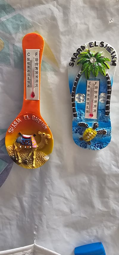 Two novelty thermometers are displayed on a white textured surface. The thermometer on the left is orange and shaped like a spoon, with a camel figurine and desert landscape at the base. Text above the camel reads "SHARM EL SHEIKH". The thermometer on the right is shaped like a flip-flop, with a palm tree and a sea turtle at the base. Text above the palm tree reads "SHARM EL SHEIKH". Both thermometers have Celsius and Fahrenheit scales, with the mercury indicating a temperature between 20 and 30 degrees Celsius. The image does not depict Karshi City, Uzbekistan.