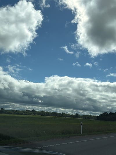 The image is a landscape photograph taken from a vehicle, showing a split view between the sky and the ground. On the left side of the image, the sky is a vibrant blue, filled with a variety of clouds. Some clouds are large, white, and puffy, while others are thinner and wispy. A large, dark cloud bank dominates the upper left portion of the frame.
On the right side of the image, the scene transitions to the ground. A dark asphalt road runs parallel to a lush green field. The field is bordered by a dense line of trees. A white road marker with black accents stands in the field next to the road. The image appears to be taken during the daytime, with the sun casting bright light on the clouds and the field. The weather seems to be mixed, with both sunny and cloudy conditions present. The overall impression is one of a rural setting in Estonia, possibly along a highway. No people are visible in the image, and there is no visible text.