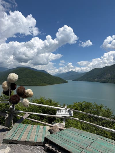 A vertical, outdoor, daytime photograph captures a scenic mountain and lake landscape in Aranisi, Georgia. In the foreground, a weathered wooden structure with peeling green paint, possibly a viewing platform or bench, is visible on the right. Attached to a rustic white railing are several fluffy, round objects, resembling hats or decorations, in shades of white and brown. A white sign with black lettering is also attached to the railing, with the visible text reading "FREE PHOTO."
Behind the railing, a vast, serene lake stretches into the distance, its calm surface reflecting the pale blue sky. Majestic, densely forested mountains rise from the opposite shore of the lake, their slopes covered in a rich green hue. The sky above is a vibrant blue, dotted with numerous large, fluffy white clouds that cast soft shadows on the landscape. The overall atmosphere is peaceful and idyllic, suggestive of a tourist viewpoint offering breathtaking natural beauty. The bright sunlight and clear skies indicate fair weather, likely during the warmer months.