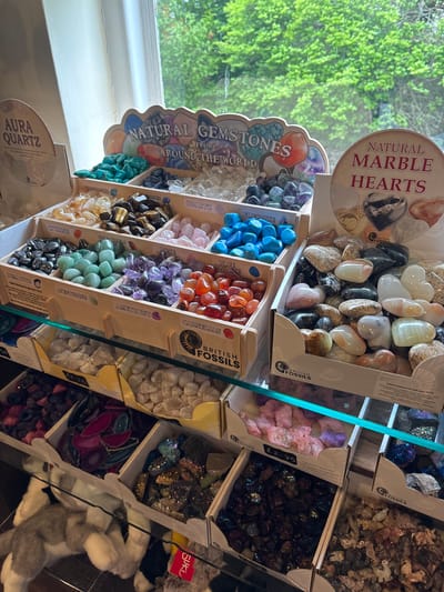 Here is a detailed description of the image:

**Setting:**

The image appears to have been taken inside a gift shop in Llanberis, United Kingdom. This is suggested by the range of items on display, the style of the signage, and the general retail setting. The location is further suggested by the "BRITISH FOSSILS" branded packaging seen in the boxes.

**Subjects and Objects:**

*   **Gemstones and Minerals:** The main focus is a display of various tumbled gemstones and minerals arranged in labeled wooden boxes or containers. Specific types include:
    *   Aura Quartz
    *   Citrine
    *   Hematite
    *   Aventurine
    *   Amethyst
    *   Carnelian
    *   Rose Quartz
    *   Quartz
    *   Fluorite
    *   Golden Tiger's Eye
*   **Marble Hearts:** A display of polished natural marble hearts in various colors.
*   **Signage:** Various signs label the different types of stones and promote the "Natural Gemstones From Around the World." The "BRITISH FOSSILS" branding is also prominent.
*   **Packaging:** The gemstones are contained in cardboard boxes with distinctive "BRITISH FOSSILS" branding.
*   **Glass Shelf:** A vertical sheet of glass acts as a barrier between the viewer and some of the gemstones.
*   **Plush Toy:** A plush toy with a yellow label and the name "SUKI" on it is visible in the lower right.
*   **Window:** A window with a view of green foliage outside is visible in the background.

**Scene:**

The scene depicts a corner of a gift shop, likely targeting tourists or rock and mineral enthusiasts. The emphasis is on displaying a variety of gemstones in an appealing and informative manner. The "BRITISH FOSSILS" branding suggests a focus on local or British-sourced items.