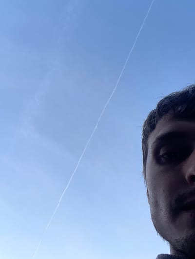 The image is a close-up, low-angle shot of the right side of a young man's face, cropped at the cheekbone and forehead. He has dark hair and a light mustache. His eye is visible, looking upwards. The background is a clear, pale blue sky with a faint contrail stretching diagonally from the lower left to the upper right of the frame. The lighting suggests it is daytime, possibly early morning or late afternoon due to the soft quality of the light. There are no discernible landmarks or indications of a specific location within the city of Genk, Belgium. The image appears to be a selfie taken outdoors.