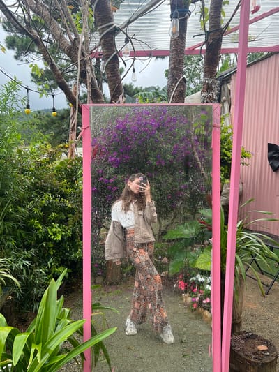 The scene is set outdoors in Đưng K'Si, Vietnam, centered on a tall, rectangular mirror with a bright pink frame. A young woman, reflected in the mirror, is capturing a selfie with a dark-colored smartphone held in her right hand. She has long, dark brown hair and is dressed in a white t-shirt, a light brown jacket draped over her left shoulder, wide-leg patterned trousers, and light-colored athletic shoes. The mirror's surface is visibly wet with water droplets, reflecting a dense bush of purple flowers immediately behind the woman. To the left of the mirror, lush green foliage and trees are present, along with a string of yellow-domed lights. To the right, a pink corrugated metal wall features a black bow decoration. Above the mirror, several tree trunks are adorned with Edison-style string lights, and a clear corrugated plastic roof structure is visible. The ground below appears to be wet gravel under an overcast sky, indicating recent precipitation.