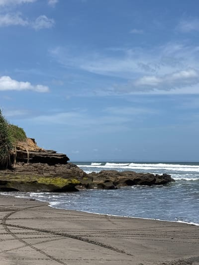 The image depicts a coastal scene in Kerambitan, Indonesia. The sky is blue with wispy clouds. The ocean is visible with small waves breaking near the shore. Rocky cliffs with some vegetation are located along the edge of the beach. The sand is dark in color with several tire tracks visible. There are no people depicted in the scene.