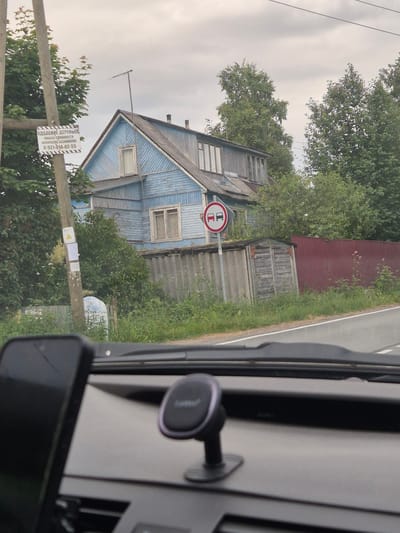 In Mashezero, Russia, an outdoor scene captured from within a vehicle shows a residential area with a rustic blue wooden house. In front of the house, a traffic sign prohibiting overtaking is visible. The house, surrounded by overgrown vegetation, features a weathered appearance with visible wood planks and multiple windows. To the left of the house, a wooden utility pole stands with several signs attached, one with text and a warning triangle. A red metal fence runs along the right side of the property, and a small, dilapidated wooden shed is situated near the road. The foreground of the image shows the interior of the vehicle, including a dashboard with a phone mount. The sky is overcast, suggesting a cloudy day.