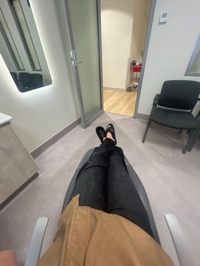 The image shows a person sitting in a waiting room. The person is wearing black pants and black sandals, and a tan jacket. The room is mostly white and grey, with a door partially visible in the background. There is a black chair to the left, and a mirror on the wall. The floor appears to be light grey. A counter or sink is visible in the lower left corner. The perspective is from above the person's legs.