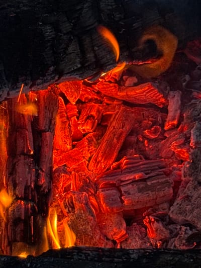 The image depicts a close-up view of a bonfire with glowing embers and flames. The dominant colors are red, orange, and black, highlighting the intense heat and texture of burning wood. The scene is set in Imelevo, Russia, and shows a close-up of burning logs, embers, and flames. There are no people or animals visible in the image. The environment appears to be outdoors, likely a natural setting given the context of a bonfire. The focus is on the dynamic and fiery nature of the combustion process, with the wood elements showing charred surfaces and the embers emitting a strong red glow.
