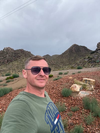 A medium shot captures a man in a green t-shirt and aviator sunglasses posing for a selfie. He is standing outdoors, with a natural landscape of rocky hills and sparse vegetation behind him. The ground is covered in reddish-brown gravel and small rocks, with a few larger boulders scattered around. The sky is overcast with a gray, cloudy ceiling. Some of the boulders have graffiti on them. The overall impression is of a rugged, arid environment. The man's face is partially obscured by his sunglasses, but he appears to be smiling slightly. The scene is static, with no apparent action or interaction occurring. The location is identified as Etholen, United States.