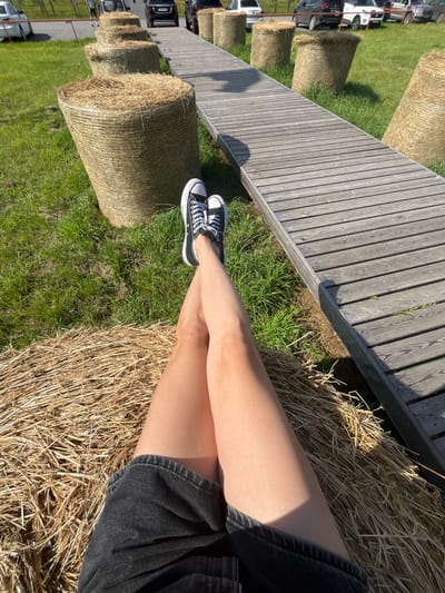 The image shows a person's legs and feet, wearing black Converse sneakers and a dark denim skirt, resting on a bale of hay. The hay bale is positioned on a grassy area with several other hay bales and a wooden boardwalk in the background. Cars are visible parked on a road further in the background, suggesting an outdoor recreational or event space. The sun is casting shadows, indicating it is daytime. The location is identified as Akhlebaikha (Kovriginskii S/S), Russia.