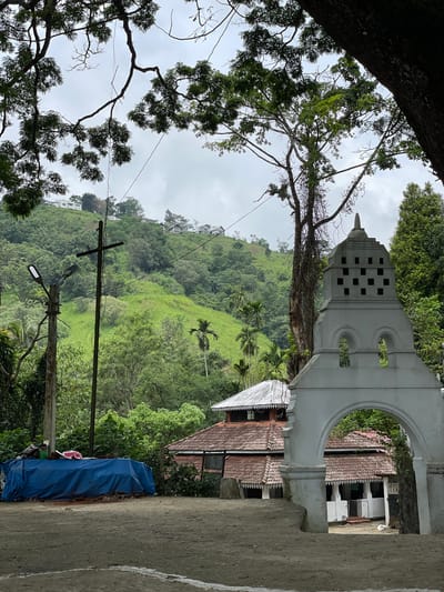 The image depicts a lush hillside in Kumbalwela, Sri Lanka, framed by a large tree on the left and a white arched building structure on the right. The hillside is densely vegetated with various trees and foliage, and several small buildings or houses are visible higher up the slope. An electrical pole with wires and a street light stands in the foreground, slightly off-center. A blue tarp is partially visible in the lower right corner, alongside a concrete wall or sidewalk. The white building features a distinctive tower with small square openings. The sky is overcast with grey clouds.