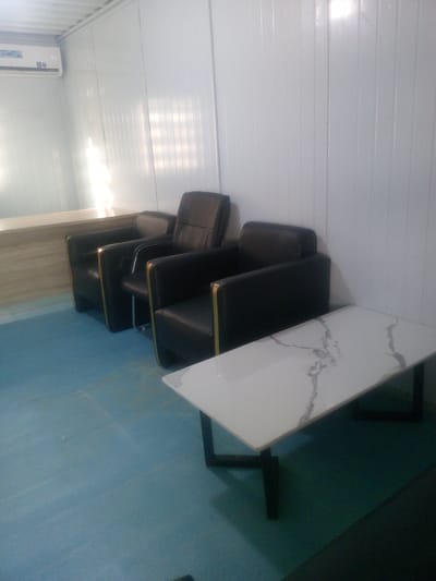 This media file depicts an indoor waiting area with a minimalist design. The scene features three black leather armchairs with gold trim, arranged along a white paneled wall. In front of the chairs, a rectangular coffee table with a white marble-patterned top and black metal legs is placed. To the left, a portion of a light-colored, possibly wooden, counter or reception desk is visible. The floor is a solid light blue color. Above the counter, a white air conditioning unit is mounted on the wall. The room appears to be well-lit, with a bright, possibly natural light source reflecting off the white walls. The overall impression is one of a clean, modern, and potentially professional waiting space. There are no people visible in the image. The time of day and weather are not discernible. The location cues suggest an indoor environment, possibly an office, clinic, or reception area in Indo, Nigeria. No specific activity or event is taking place, as the scene is static. There is text visible on the air conditioning unit, but it is too small and blurry to read.