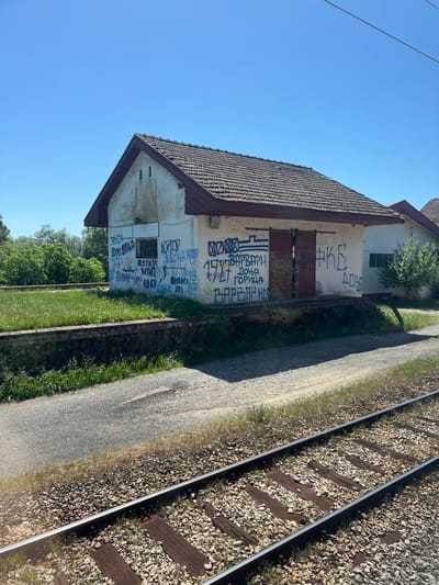 An exterior shot shows a two-story building covered in graffiti in Bistrice, Montenegro. The building, with a roof of terracotta tiles and white walls, stands adjacent to a railway track that runs alongside a dirt road. Graffiti is extensively spread on the building's walls. The sky is bright blue. Green foliage is visible at the base of the building. Overhead electric lines are visible in the upper left of the image.