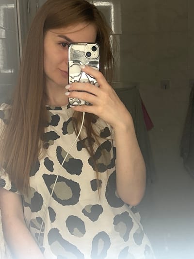 A young woman with long, auburn hair is taking a selfie in a mirror. She is wearing a tan and black leopard print t-shirt. Her right hand, with freshly manicured white nails, is holding up a silver phone with a skull-shaped case, obscuring her face. A white earphone cord is visible, running from the phone down her shirt. The background is dimly lit and appears to be a bathroom, with a mirror reflecting the scene and some hanging clothes in the distance. The image does not provide specific details to place it in the city of Нижние Муллы, Russia.
