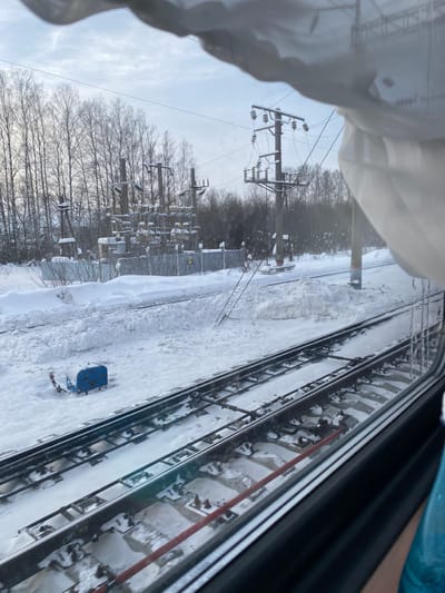The scene depicts a snow-covered railway environment, observed from a train window in what is consistent with the city of Yar, Russia. In the foreground, two parallel railway tracks are visible, partially covered by snow, with dark sleepers discernable between the rails. A dark blue utility box with a red lever is situated on the snow-covered ground adjacent to the tracks. Beyond substantial snow drifts and a chain-link fence, an electrical substation is present, consisting of multiple concrete poles supporting various insulators and electrical equipment, all heavily coated in snow. Several utility poles with associated power lines stand in the midground; one pole appears to be leaning significantly, with wires extending towards the ground. The background features a dense line of bare deciduous trees, likely birches, covered in snow, under an overcast, light grey sky. No individuals or discernible actions are depicted.