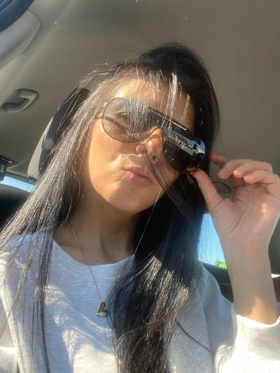 An adult female is depicted in a close-up portrait, situated within the interior of a vehicle, identifiable by the visible headliner in the background. She possesses long, dark hair, which is illuminated by direct sunlight, casting highlights across her face and hair. She wears oversized, dark-tinted aviator-style sunglasses, obscuring her eyes. Her lips are slightly pursed in a "kissing" pose. Her right hand is positioned at the right temple of her sunglasses, lightly grasping the frame. She is attired in a white, long-sleeved shirt, and a gold-colored necklace with a heart-shaped pendant is visible around her neck. Reflections within the sunglasses' lenses show an outdoor scene, including a clear blue sky and what appears to be a multi-story building with multiple windows, possibly a commercial or residential structure, along with indistinct vehicles or environmental features. The precise location within Bensheim, Germany, is not definitively ascertainable from the reflections.